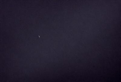 The primary subject of the image is a crescent moon, which is visible in the sky. The setting is a dark, cloudy night with a black background. The visual style is a photo, capturing the moon in its natural environment. The colors and mood of the image are predominantly dark and moody, with the moon being the only bright spot in the sky.