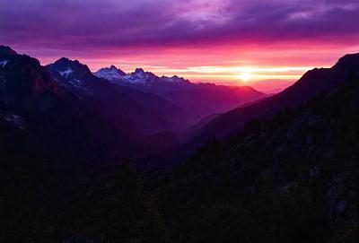 The primary subject of the image is a beautiful mountain range with a sunset in the background. The setting is a mountainous area with a purple sky, and the visual style is a photo. The colors and mood of the image are serene and picturesque, with the sunset casting a warm glow over the mountains.