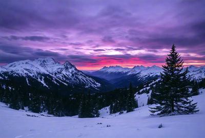 The primary subject of the image is a beautiful mountain range with a purple sky in the background. The setting is a snowy mountain top, and the visual style is a photo. The colors and mood of the image are serene and breathtaking, with the purple sky adding a touch of magic to the scene.