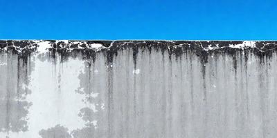 The primary subject of the image is a concrete wall with a peeling paint surface. The setting is an outdoor environment, with a blue sky visible in the background. The visual style is a photo, capturing the peeling paint on the concrete wall. The colors in the image are predominantly blue and grey, creating a somewhat dull and aged atmosphere.