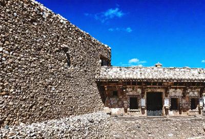 The primary subject of the image is a large, old stone building with a rustic appearance. The building is made of stone and has a unique, textured appearance. The setting is outdoors, with a clear blue sky visible in the background. The visual style of the image is a photo, capturing the details and textures of the stone building. The colors in the image are predominantly shades of blue and gray, creating a calm and serene atmosphere.