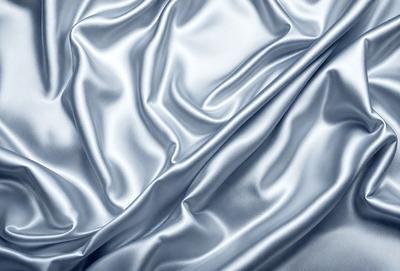 The primary subject of the image is a piece of shiny, silky fabric. The setting is a white background, which provides a clean and minimalist appearance. The visual style is a close-up of the fabric, showcasing its texture and reflective quality. The colors and mood of the image are predominantly silver and white, creating a sense of elegance and sophistication.
