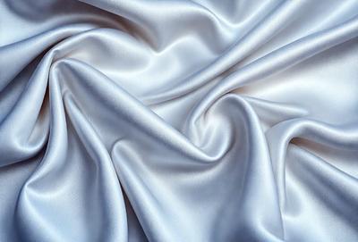 The primary subject of the image is a white fabric, possibly a sheet or a curtain, with a silky texture. The setting is a white background, which provides a clean and minimalist appearance. The visual style is a close-up of the fabric, showcasing its details and texture. The colors and mood of the image are predominantly white, giving it a clean, serene, and elegant atmosphere.