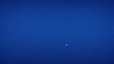 The primary subject of the image is the moon, which is seen in the sky. The setting is a vast, dark blue sky, with no clouds or other objects visible. The visual style is a black and white photo, which adds a sense of timelessness and simplicity to the scene. The colors and mood of the image are predominantly dark and moody, with the moon being the only bright spot in the sky.