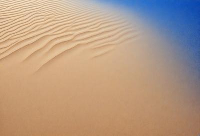The image features a sandy beach with a sandy surface. The primary subject is a close-up of the sand, showcasing its texture and appearance. The setting is a beach environment, and the visual style is a natural, realistic representation of the sand. The colors and mood of the image are predominantly warm and earthy, with the sand being the main focus of the scene.