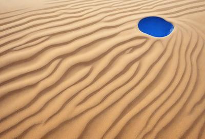 The primary subject of the image is a sandy surface with a blue hole in the middle. The setting is a desert-like environment, with sand covering the ground. The visual style is a close-up of the sandy surface, showcasing the intricate patterns and textures of the sand. The colors and mood of the image are predominantly earthy tones, with the blue hole adding a touch of contrast and interest to the scene.