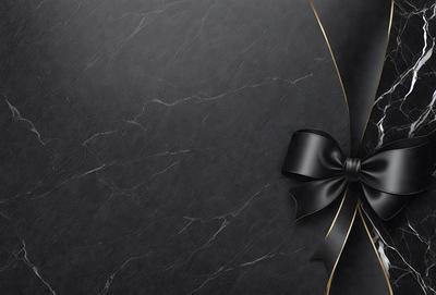 The primary subject of the image is a black bow, which is prominently displayed on a black background. The setting appears to be a marble wall, adding a sense of sophistication and elegance to the scene. The visual style of the image is a close-up of the bow, emphasizing its details and texture. The colors and mood of the image are predominantly black, creating a dramatic and striking appearance.
