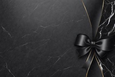 The primary subject of the image is a black bow, which is prominently displayed on a black background. The setting appears to be a marble surface, which adds a sense of sophistication and elegance to the scene. The visual style of the image is a close-up of the bow, emphasizing its details and texture. The colors and mood of the image are predominantly black, creating a sense of formality and refinement.