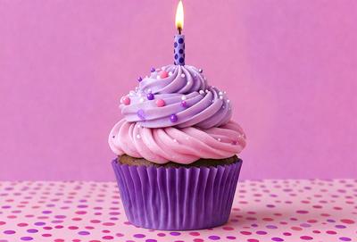 The primary subject of the image is a cupcake with a candle on it. The cupcake is purple and has white frosting. The setting is a pink background, which adds a warm and inviting atmosphere to the scene. The visual style is a close-up of the cupcake, highlighting its details and making it the focal point of the image. The colors and mood convey a sense of celebration and joy, as the cupcake is often associated with special occasions and treats.