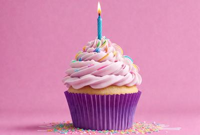 The primary subject of the image is a cupcake with a candle on top, placed on a pink surface. The cupcake is decorated with pink frosting and sprinkles, giving it a colorful and festive appearance. The setting appears to be a celebration or a special occasion, as the cupcake is adorned with a candle, suggesting that it might be a birthday or another event. The visual style of the image is a close-up of the cupcake, highlight