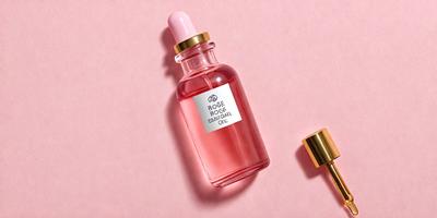 The primary subject of the image is a bottle of Rose Bogue Oil, which is placed on a pink surface. The setting is a close-up of the bottle, showcasing its pink color and the brand name. The visual style is a realistic photograph, capturing the details of the bottle and its surroundings. The colors and mood of the image are predominantly pink, creating a warm and inviting atmosphere.