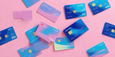 The primary subject of the image is a collection of various credit cards. These cards are spread out across the scene, with some placed on a pink surface. The cards come in different colors and designs, showcasing a diverse range of options for consumers. The visual style of the image is a close-up of the credit cards, highlighting their details and making them the main focus of the scene. The colors and mood of the image are vibrant and lively, with the pink surface adding a touch of warmth