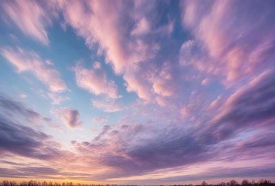 The primary subject of the image is a beautiful sunset with a cloudy sky. The setting is a field with trees in the background, creating a serene and picturesque scene. The visual style is a photo, capturing the natural beauty of the sunset and the cloudy sky. The colors and mood of the image are predominantly warm and calming, with the sunset casting a warm glow over the landscape.