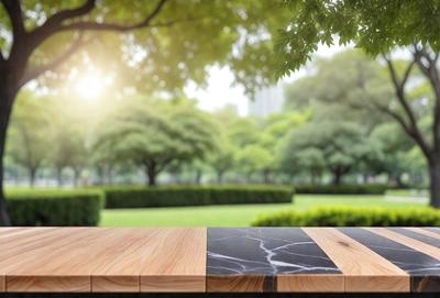 The primary subject of the image is a wooden table with a black marble top. The table is placed in a park setting, surrounded by trees and grass. The visual style of the image is a photo, capturing the natural environment and the table's design. The colors and mood of the image are predominantly green and serene, as the park is filled with lush greenery and the wooden table adds a warm, natural touch to the scene.