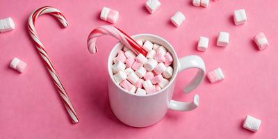 The primary subject of the image is a cup filled with marshmallows and candy canes. The cup is placed on a pink surface, which adds a festive touch to the scene. The visual style of the image is a close-up, showcasing the details of the marshmallows and candy canes. The colors in the image are predominantly pink, which creates a warm and inviting atmosphere.