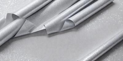The primary subject of the image is a roll of silver paper, which is the main object in the scene. The paper is sitting on a table, and it appears to be a patterned material. The setting is a table, and the visual style is a photo. The colors and mood of the image are predominantly silver and white, giving it a clean and elegant appearance.