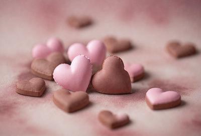 The primary subject of the image is a collection of heart-shaped chocolates. The setting is a pink surface, which adds a warm and romantic atmosphere to the scene. The visual style is a close-up of the chocolates, showcasing their details and textures. The colors are predominantly pink, which is associated with love and affection.