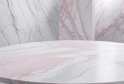 The primary subject of the image is a large, round, white marble table. The table is placed in a room with a pink marble wall, which adds a touch of color to the scene. The visual style of the image is a photo, capturing the details of the marble table and the pink marble wall. The colors and mood of the image are predominantly white and pink, creating a clean and elegant atmosphere.