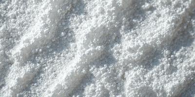 The primary subject of the image is a close-up of a white powder, likely flour or sugar, with a slightly speckled texture. The setting is a white surface, possibly a table or a countertop, where the powder is placed. The visual style appears to be a close-up of the powder, with a focus on its texture and appearance. The colors in the image are predominantly white, giving the scene a clean and simple appearance.