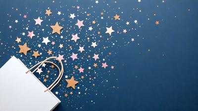The primary subject of the image is a white shopping bag with gold stars on it. The bag is placed on a blue background, which adds a contrasting color to the scene. The visual style of the image is a combination of photo and digital art, creating a unique and eye-catching design. The colors and mood of the image are predominantly white and gold, giving it a warm and inviting atmosphere.