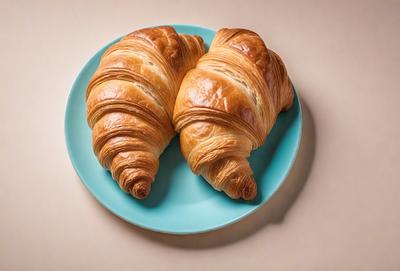 The primary subject of the image is a plate with two croissants on it. The croissants are placed on the plate in a way that they appear to be facing each other. The setting is a dining table, and the visual style is a close-up of the plate with the croissants. The colors and mood of the image are warm and inviting, as the croissants are golden and the overall atmosphere is cozy and appetizing.