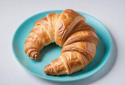 The primary subject of the image is a croissant, which is the main object, scene, or focus of the image. The croissant is placed on a blue plate, which serves as the setting or background. The visual style of the image is a photo, capturing the details and colors of the croissant. The dominant colors in the image are blue and white, creating a calm and inviting atmosphere.