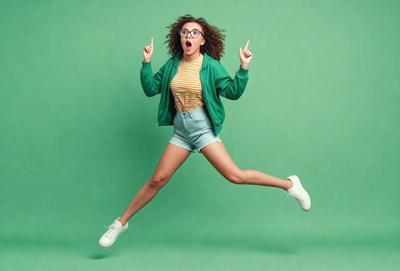 The primary subject of the image is a woman wearing a green jacket and glasses, who is jumping in the air with her arms raised. She appears to be celebrating or expressing excitement. The setting is a green background, which adds a vibrant and lively atmosphere to the scene. The visual style is a photo, capturing the woman's energetic moment. The colors and mood of the image are lively and joyful, as the woman's actions and the green background create a sense of