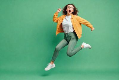 The primary subject of the image is a woman wearing glasses and a yellow jacket, who is jumping in the air with her arms raised. She is the main focus of the scene. The setting is a green background, which adds a vibrant and lively atmosphere to the image. The visual style is a photo, capturing the woman's action and expression. The colors and mood of the image are energetic and joyful, as the woman appears to be having a great time while jumping.