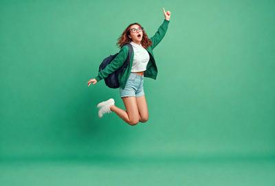The primary subject of the image is a woman wearing a green jacket and glasses, who is jumping in the air with her arms raised. She is holding a backpack and appears to be celebrating or enjoying herself. The setting is a green background, which adds a vibrant and lively atmosphere to the scene. The visual style is a photo, capturing the woman's joyful moment. The colors and mood of the image are energetic and cheerful, with the woman's green jacket