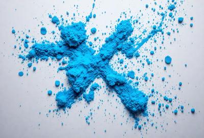 The primary subject of the image is a blue powder or dust, which is scattered across a white surface. The setting appears to be a laboratory or a workspace, as the blue powder is being analyzed or studied. The visual style of the image is a close-up of the blue powder, showcasing its texture and color. The dominant colors in the image are blue and white, creating a calm and focused atmosphere.