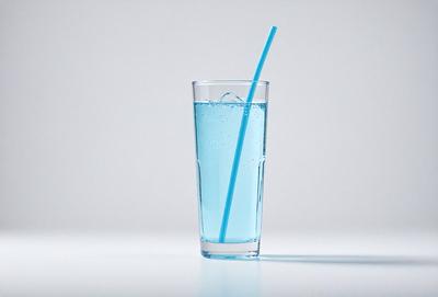 The primary subject of the image is a blue drink in a clear glass. The drink is filled with bubbles, indicating that it is carbonated. The glass is placed on a white surface, which serves as the setting or background of the image. The visual style of the image is a photo, capturing the details of the drink and the glass. The dominant colors in the image are blue and white, creating a calm and refreshing atmosphere.