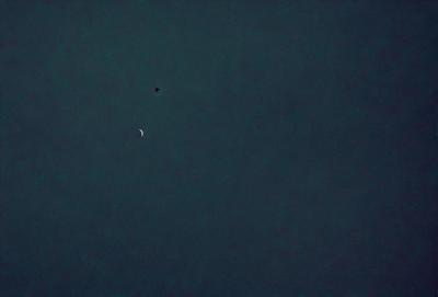 The primary subject of the image is a moon, which is visible in the sky. The setting is a dark, cloudy night with a blue sky. The visual style is a photo, capturing the moon and its surroundings. The colors and mood of the image are predominantly dark and moody, with the moon being the main source of light in the scene.