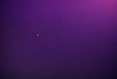 The primary subject of the image is a moon, which is visible in the sky. The setting is a dark, purple sky, which creates a dramatic and moody atmosphere. The visual style of the image is a photo, capturing the moon and the night sky. The colors and mood of the image are predominantly dark and mysterious, with the purple sky adding a sense of depth and intensity to the scene.
