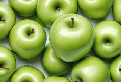 The primary subject of the image is a group of green apples. They are arranged on a white surface, which could be a table or a counter. The apples are placed in various positions, with some closer to the front and others further back. The visual style of the image is a close-up of the apples, showcasing their vibrant green color and natural texture. The overall atmosphere of the image is fresh and inviting, with the green apples being the main focus of the scene.