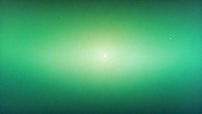 The primary subject of the image is a bright, glowing sun. The sun is positioned in the center of the image, surrounded by a dark, green background. The visual style of the image is a photo, capturing the sun's light and the contrasting darkness of the background. The colors and mood of the image are predominantly dark and moody, with the sun being the only source of light in the scene.