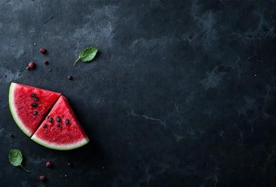 The primary subject of the image is a slice of watermelon. The watermelon slice is placed on a black surface, which could be a table or a countertop. The setting appears to be indoors, and the visual style is a close-up of the watermelon slice. The colors in the image are predominantly black and red, giving the image a bold and vibrant appearance.