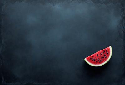 The primary subject of the image is a slice of watermelon, which is placed on a black surface. The watermelon slice is the main focus of the image, and it is presented in a close-up view. The setting or background consists of a black surface, which contrasts with the watermelon slice. The visual style of the image is a close-up, and the colors are predominantly black and white, giving the image a simple and minimalist appearance.