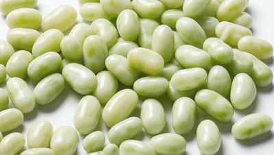 The primary subject of the image is a pile of green beans. The beans are arranged in a visually appealing manner, with some beans overlapping others. The setting is a white surface, which provides a clean and uncluttered backdrop for the beans. The visual style of the image is a close-up of the beans, showcasing their texture and details. The colors and mood of the image are predominantly green, which gives the scene a fresh and natural feel.