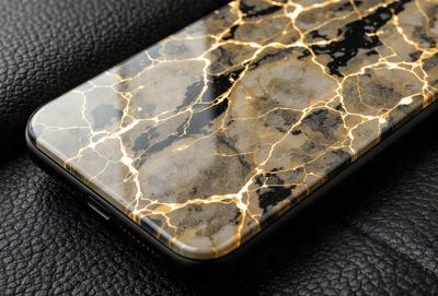 The primary subject of the image is a cell phone with a marble-like pattern on its back. The setting is a leather surface, and the visual style appears to be a close-up of the cell phone. The colors and mood of the image are predominantly black and gold, giving it a luxurious and elegant appearance.