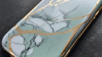The primary subject of the image is a cell phone case with a marble design. The case is made of marble and features gold accents. The setting is a table, and the visual style is a close-up of the cell phone case. The dominant colors are marble and gold, giving the case a luxurious and elegant appearance.