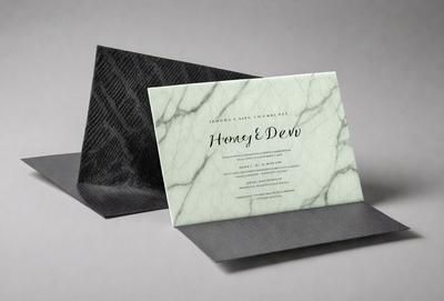 The primary subject of the image is a card, which is placed on a grey background. The card is a square-shaped invitation, possibly for a wedding or a similar event. The visual style of the image is a combination of photo and digital art, as it appears to be a real card but has a digital-like appearance. The colors and mood of the image are predominantly grey, giving it a sophisticated and elegant feel.