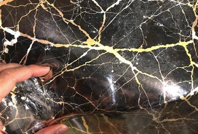 The primary subject of the image is a marble-like surface with a gold-like pattern. The setting is a room with a person holding the marble-like surface. The visual style is a close-up of the marble-like surface, showcasing its unique texture and design. The colors and mood of the image are predominantly gold and brown, giving it a warm and luxurious appearance.