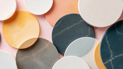 The primary subject of the image is a collection of round, white, and blue plates. The plates are arranged in a visually appealing manner, with some placed on a pink surface. The setting appears to be a studio or a workspace, as the plates are displayed in a way that showcases their unique designs. The visual style of the image is a combination of photo and digital art, creating a vibrant and artistic atmosphere. The dominant colors in the image are white, blue, and pink, which