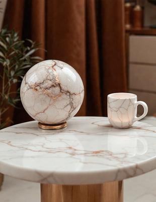 The primary subject of the image is a white marble sphere, which is placed on a table. The setting is a room with a table and a chair. The visual style is a photo, and the colors and mood are predominantly white, giving the scene a clean and elegant appearance.
