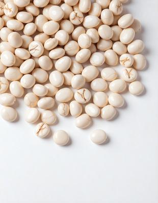 The primary subject of the image is a pile of white beans. The beans are scattered across a white surface, which could be a table or a counter. The setting appears to be a kitchen or a dining area, as the beans are placed on a table or counter. The visual style of the image is a close-up of the beans, showcasing their texture and details. The colors in the image are predominantly white, giving the scene a clean and simple appearance.