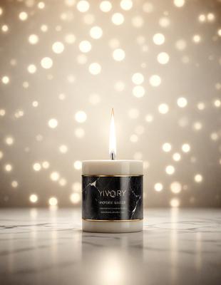 The primary subject of the image is a lit candle in a glass jar. The candle is placed on a table, and the jar is decorated with a marble pattern. The setting is a room with a patterned wall, and the visual style is a close-up of the candle and jar. The colors and mood of the image are warm and inviting, with the lit candle creating a cozy atmosphere.