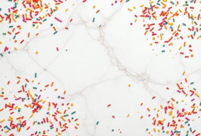 The primary subject of the image is a marble countertop with a variety of sprinkles scattered across it. The setting is a kitchen or dining area, and the visual style appears to be a photo of the countertop. The colors and mood of the image are vibrant and cheerful, with the sprinkles adding a playful and fun atmosphere to the scene.