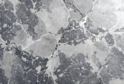 The primary subject of the image is a close-up of a marble surface with a pattern of holes and grooves. The setting is a stone wall, and the visual style is a photo. The dominant colors are gray and black, creating a moody atmosphere.