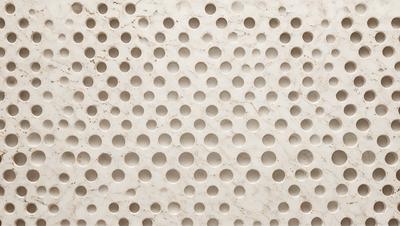 The primary subject of the image is a white marble wall with a pattern of holes in it. The setting is a room with a white marble surface, which gives the room a clean and elegant appearance. The visual style of the image is a close-up of the marble wall, showcasing the intricate pattern of holes. The colors and mood of the image are predominantly white, giving it a calm and serene atmosphere.
