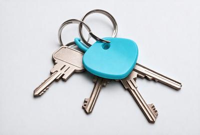 The primary subject of the image is a pair of keys. The keys are blue and silver, and they are placed on a white surface. The setting appears to be indoors, possibly in a room with a table. The visual style of the image is a close-up of the keys, emphasizing their details and colors. The overall atmosphere of the image is clean and organized, with the keys being the main focus of the scene.